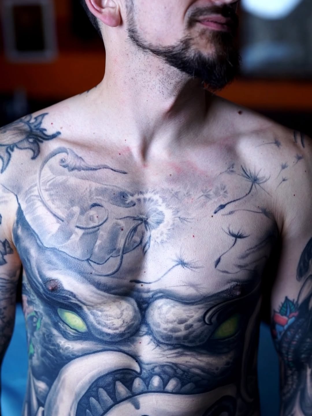 black-and-grey dandelion blowing seeds across upper chest, with an existing demon-mask stomach piece below